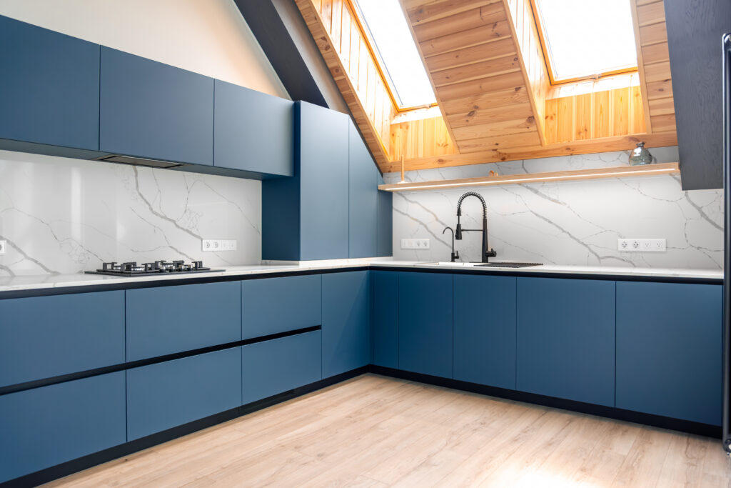 Southern Cabinets of Charleston presents a modern L-shaped kitchen in an attic with a vaulted wood-clad ceiling and skylights. The kitchen showcases matte blue, frameless cabinets with an integrated dark handle profile.