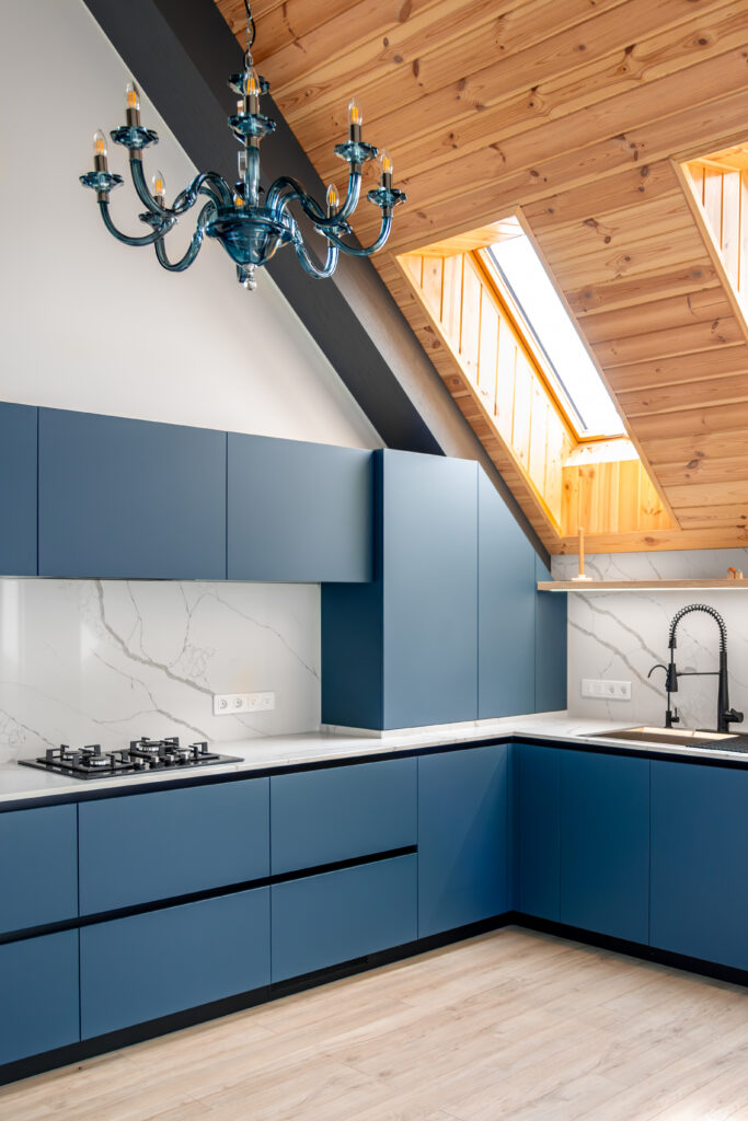 A modern loft kitchen featuring deep navy-blue two-tone cabinets, white marble backsplash and countertops, a black gas cooktop, matte black faucet, and a statement blue glass chandelier. Natural light pours in through a skylight window, highlighting the warm wood-paneled vaulted ceiling — a perfect showcase of custom two-tone kitchen cabinet design by Southern Cabinets of Charleston.