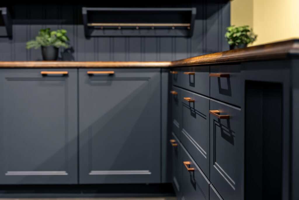 Close-up of matte charcoal grey kitchen cabinets with copper drawer pulls and a warm wood countertop, featuring layered paneling details and greenery accents.