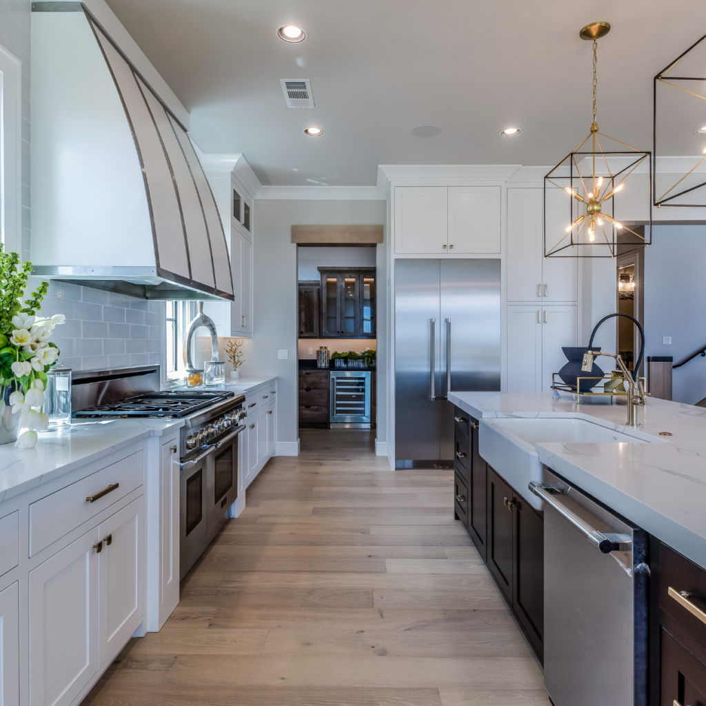 Modern chef's galley kitchen layout featuring white perimeter cabinets, a custom metallic range hood, a dark island with an apron sink, and light oak flooring leading to a butler's pantry.