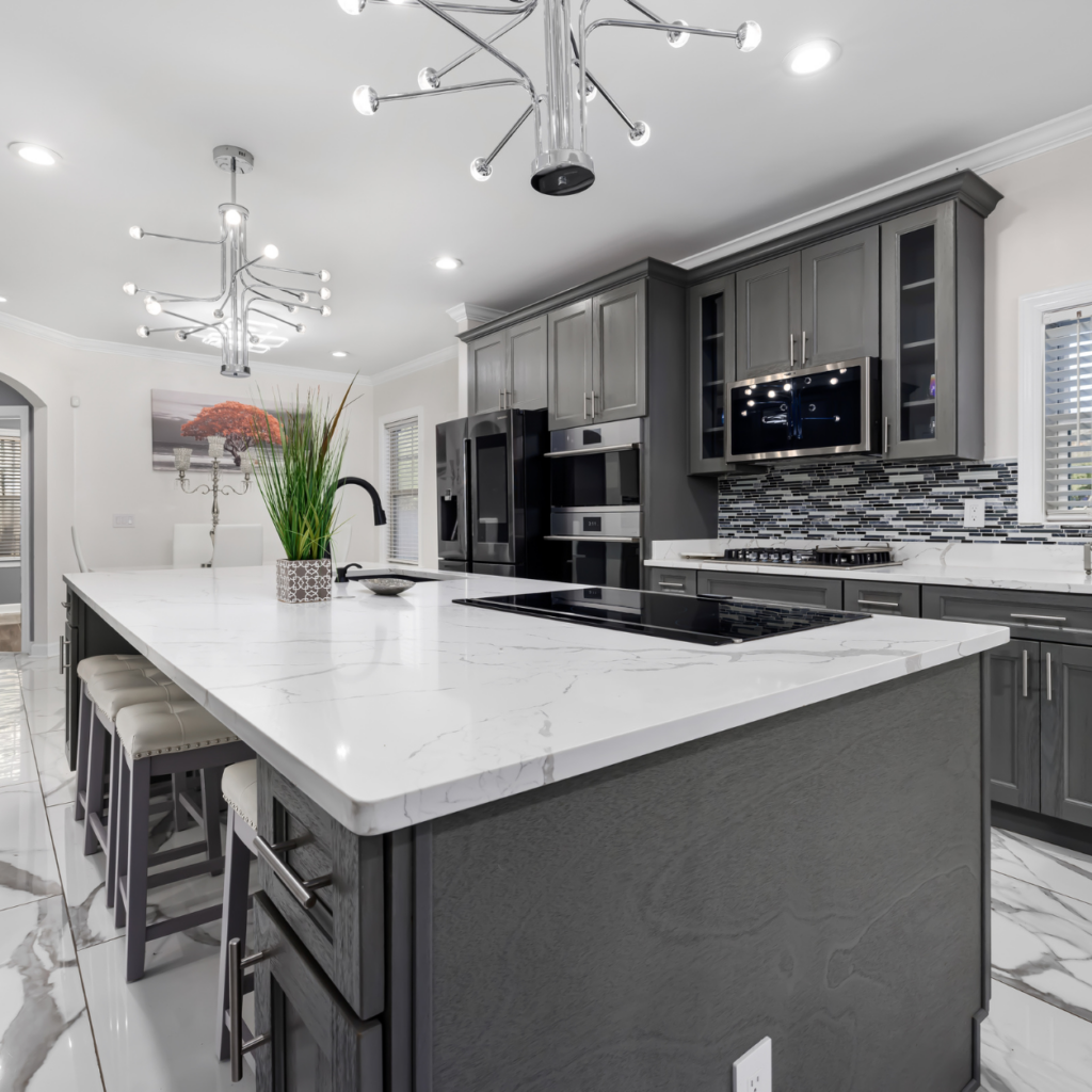 Contemporary dark gray kitchen cabinet layout featuring a large white quartz island with an integrated cooktop, modern chandelier, and polished marble flooring.